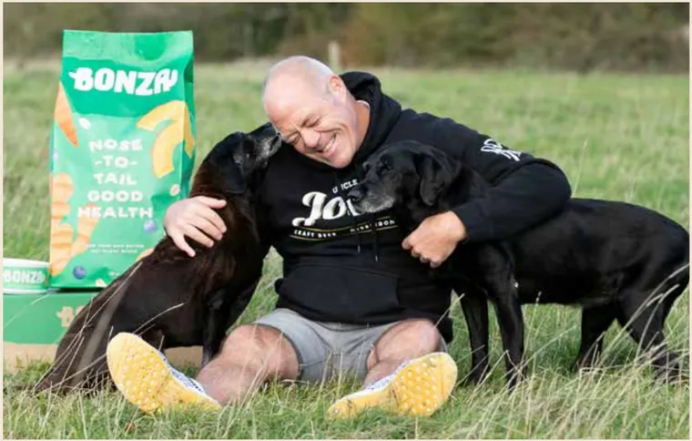 Glendon Lloyd, Founder and CEO of Bonza, sitting in a field with two black Labradors
